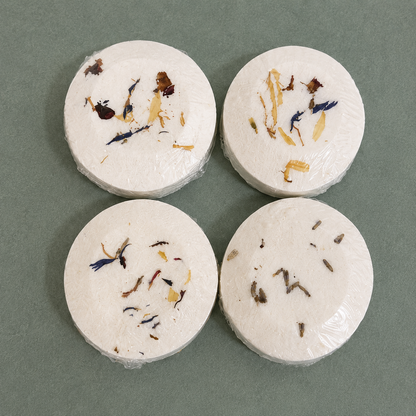 Four handcrafted aromatherapy shower steamers with dried botanicals on a sage green background, including Lavender, Balance, Headspace, and Deep Reset.